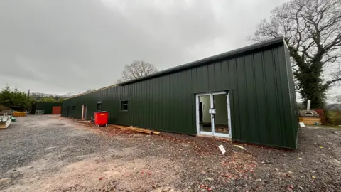 ELLEN KNIGHT/BBC A large, single-storey building painted dark green, with dark framed windows and a pair of glass doors at the front. The sky is grey and overcast, and the ground around the building is covered in gravel and mud. 