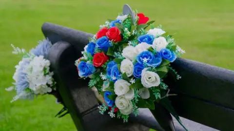 Memorial flowers are placed on a seat at a public park following the death of a person in a road accident. The flowers are in different colours, and the bench is placed near grass.