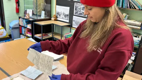 A young woman wearing a burgundy-coloured sweater marked in white letters with "English Heritage". She has long dark blonde hair under a red beanie hat and is looking at an old letter, which she is holding with blue gloves.