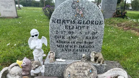 A gravestone, with the name "Kurtis George Elliott" on it, and various ornaments resting at the base