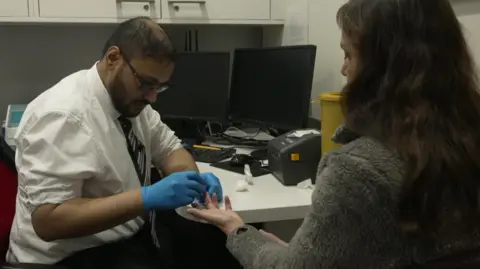 A man wearing a white shirt, tie and blue gloves takes a finger-prick blood sample from a woman seated across a desk, with computer monitors and medical equipment visible in the room.