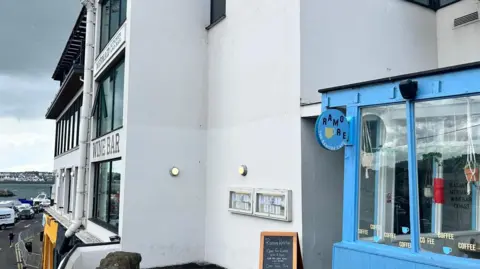 the exterior of the ramore wine bar and ramore cafe in Portrush. The images shows the cafe in the foreground. It is glass fronted and has blue window frames. The wine bar is seen on the far left and is also glass fronted and painted white
