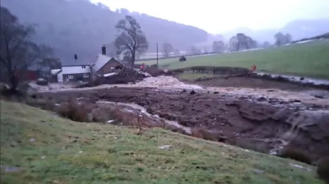 Emyr Owens A brick farmhouse pictured from a distance with tonnes of brown silt covering the yard in front of it. A pile of branches and fallen trees can also be seen by the farmhouse.