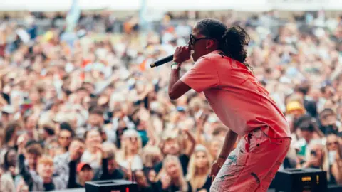Amy Fern Ms Dynamite, in T-shirt and trousers, is on stage, holding the mic to her mouth. A big crowd is seen behind her with blurred faces.