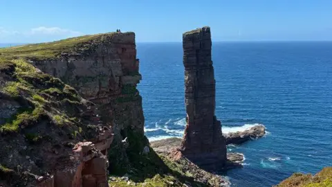 Two figures sit on a cliffside overlooking the Old Man of Hoy, an iconic sea stack in Orkney.