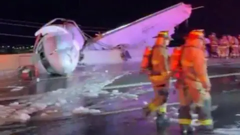 Wreckage of jet with white fusilage is lying on the motorway and two firefighters are in the foreground walking past.