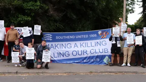 Getty Images A group of men and boys holding placards urging Ron Martin to leave Southend United. They are standing either side of a large blue and white flag that accuses Martin of killing the club.