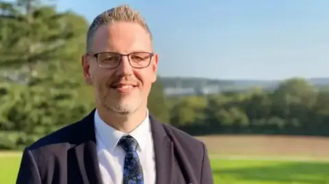Campion is standing outside with fields behind him. He is wearing a dark suit and tie and a white shirt with glasses. He is smiling at the camera and the sun is shining.
