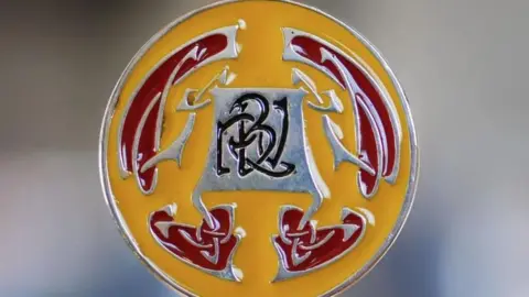CULTURE VANNIN The RBV medal which is a yellow and red disc, with a Celtic design in the style of Archibald Knox. The letters RBV are engraved in black on a silver background in the middle.