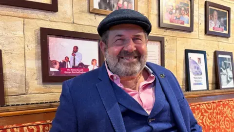 Giovanni Malacrino is pictured sat in his restaurant with a wall of celebrity photos behind him and wearing a blue three piece suit, pink shirt and flat cap and gold chain.