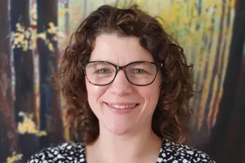 Professor Sarah Singer A woman is staring outwards and smiling. Just her head and shoulders can be seen. She has brown curly hair that comes to her shoulders. She is wearing brown and black glasses. Behind her is a wall that has some decoration on it that looks like a forest that's been painted in brown and yellow colours. 