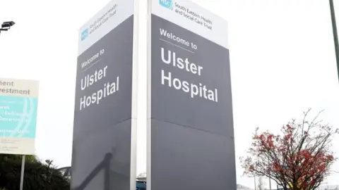A large grey sign with 'Ulster Hospital' written on it in grey writing. At the top there is a white band with 'South Eastern Health and Social Care Trust' written on it. Next to it is tree with red leaves and another sign post.