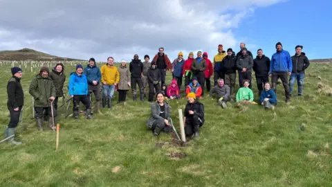 A group of volunteers who plant trees stand for a photo around the final stump of tree they planted.