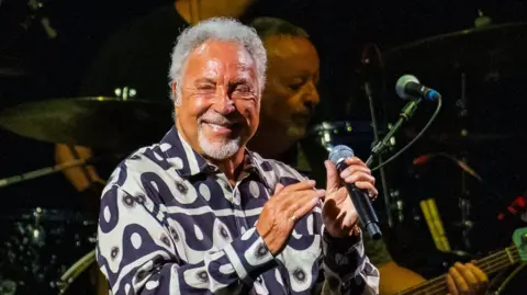 Getty Images Tom Jones holding a microphone and smiling. He is where a Black and White shirt with circular patterns on it. Behind him a guitarist and set of drums.