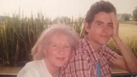 Johnny's Happy Place An old photo of a woman in a white shirt with her head resting on the shoulder if a man in a red and white chequered shirt. In the background there is a field. 