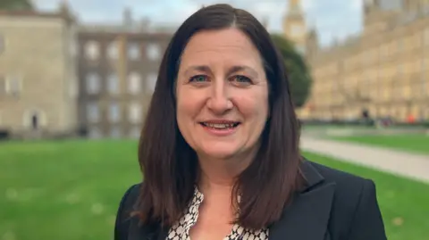 A brown-haired woman wearing a black blazer, standing on a lawn with the Palace of Westminster over her left shoulder