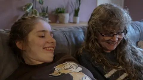 Bethany Turner and Debbie Crowson sitting on a sofa in a living room. They are smiling and looking at an electronic tablet, which is not shown in the picture.