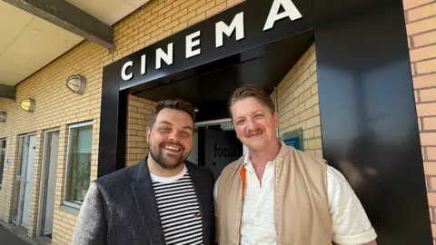 Chris Allan who is wearing a black and white striped T-shirt and a grey patterned blazer stands next to Sunderland filmmaker John Lee Taggart, who is wearing a white buttoned shirt and a beige-coloured gilet, outside the cinema at the David Puttnam Media Centre. There is an entrance where there is a black surround and the words 'CINEMA' in white lettering.