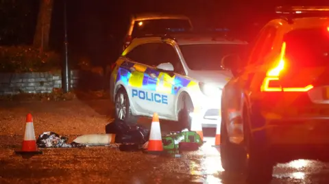 Two police cars are parked in a street, next to a few objects on the ground which have been cordoned off with traffic cones.