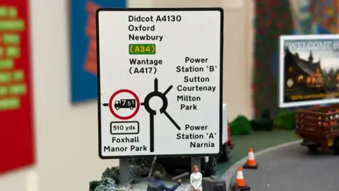 A miniature model version of the road sign in the previous image showing a roundabout. Among the locations appearing on the sign is "Narnia".