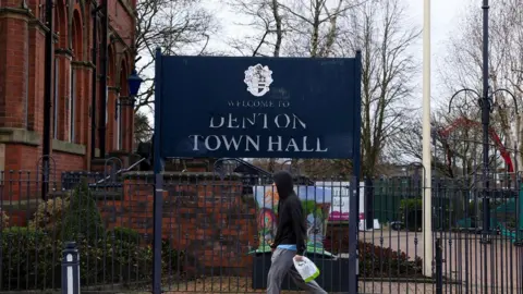 A big blue sign says Welcome to Denton Town Hall outside a redbrick historic building. A man in a hooded top with carrier bag walks past it.