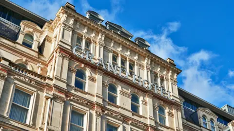 Image shows the centre and top of the Grand Hotel - it has a cream coloured stone facade and a sign in large letters reads Grand Hotel 