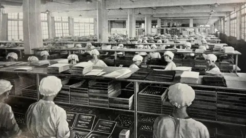 Hull History Centre An old photo, in black and white. It shows a group of about 40 factory workers all wearing white bonnets and white aprons. They are all split into rows in a factory, and are standing at production lines. In the forefront of the photo, you can see staff members are arranging sweets into boxes, titled 'Needlers' 