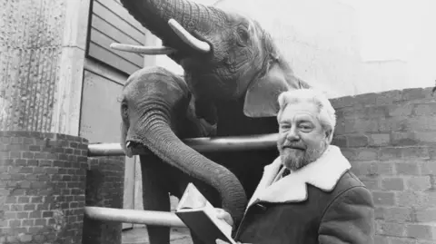 Getty Images Portrait of zookeeper and wildlife presenter Gerald Durrell with two elephants, December 1976. Durrell is wearing a cost which has sheepskin inside and on the collar, he is holding a book open and looking at the camera. Two elephants are behind him and behind a brick wall and some metal bars. One of the elephants appears to be touching Durrell's arm with its trunk.