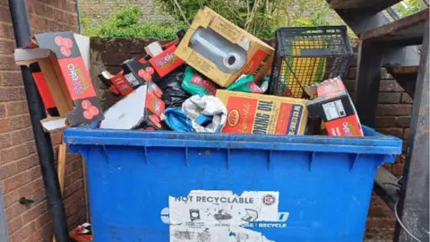 Reading Borough Council A large blue, commercial rubbish bin, piled high and overflowing with large cardboard boxes, a black bin bag and a black plastic crate. It is outside, underneath a metal staircase, with a red brick wall on the left of the image and a black plastic down pipe.