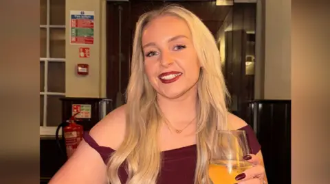 Jessica Thorpe smiling at the camera. She had long blond hair and is wearing a burgundy dress, with matching burgundy painted nails. She is holding a wine glass.