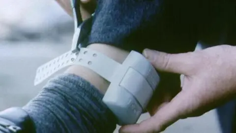 A grey ankle tag being strapped on. A strap runs through a grey box and has several holes available for different sized ankles.