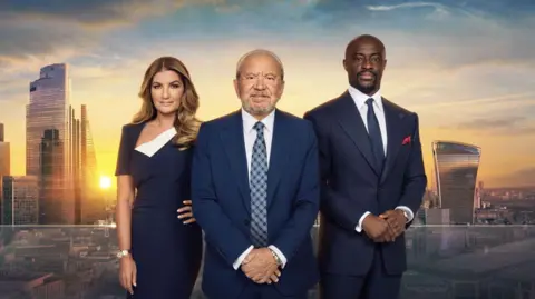 Lord Alan Sugar wears a blue suit and blue tie. He is flanked on his left by business executive Karen Brady, dressed in a blue dress, and on his right by former winner Tim Campbell, also wearing a blue suit and blue tie. They are stood in front of a backdrop of the city of London.