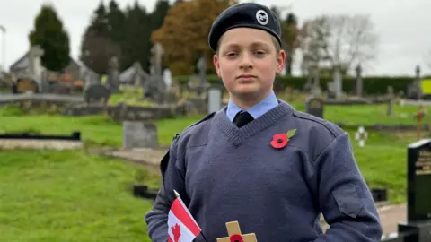 An air cadet in blue uniform, a poppy and a beret standing in a cemetery holding a small Canadian flag and a wooden remembrance cross. 