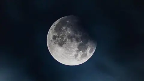 A clear picture of the white and cratered moon in a dark blue night sky.