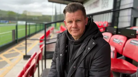 James Hughes is sitting in the seats of a football ground, looking at the camera. The seats surrounding him are red. James is wearing a black hooded jumper and a black jacket. 