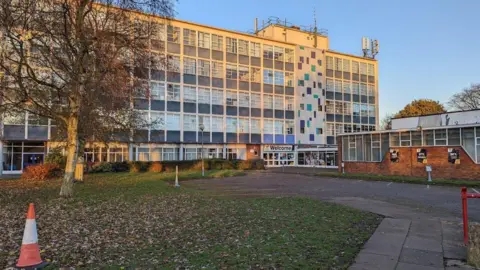A five-storey building with many windows and a welcome sign outside on the ground floor.
