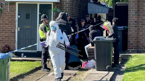 A police forensic investigator in white scrubs outside the house, with several other male and female police officers and others behind. Police tape surrounds the house