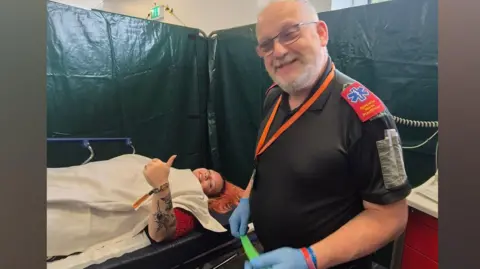 Zoe Durber-Worrall Keith has white hair and is wearing a black top with red medical shoulder pads. He is wearing blue medical gloves and is standing next to a young woman who is lying down on a medical bed under a white blanket. She has pink hair and is holding up her thumb