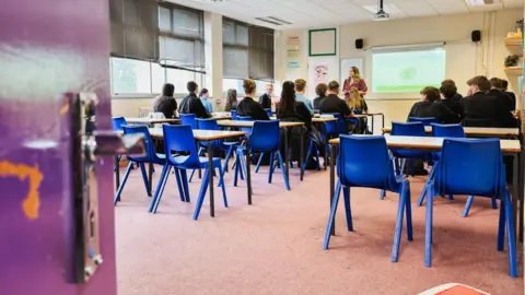 A class of pupils sat behind desks on blue chairs with their backs to the camera facing their female teacher at the front of the classroom standing next to a white board.