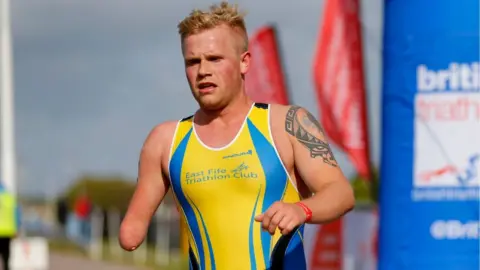 A man with blond hair in a tight yellow vest running a race. His right arm stops at the elbow, with no forearm 