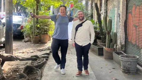 Ed Jervis Two men walk down a street during the day, with cactuses in plant pots to their left, and shutters down on buildings. Both are smiling. The man on the left is wearing white trainers, black trousers and a grey t-shirt and has his arms wide as if he is about to hug someone. The man on the right, who has dark hair and bear, wears brown shoes, black trousers and a cream long-sleeved shirt.