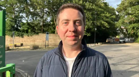 A man wearing a dark blue quilted zip-up jacket and a light-coloured top stands on a quiet road lined with mature green trees.