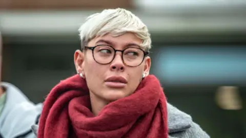 A woman with short blonde hair and glasses, wearing a grey court and red scarf