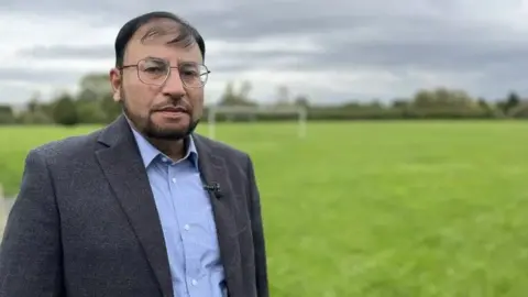 Gabby Colenso/BBC Dr Shakoor is standing on a grass field. He is wearing a grey blazer and light blue shirt. He has short dark hair and a beard and is wearing gold-rimmed glasses.