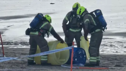 Firefighters wearing protective gear examine a blue drum on a beach. They are holding yellow drums next to the blue drum.