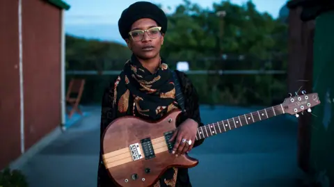Bel Odenyo stands outdoors holding a wooden electric guitar, wearing glasses, a dark hat, and a patterned scarf, centered in the frame.
 The scene is shot at dusk with a blurred background of buildings and trees, creating a calm, portrait-style composition.