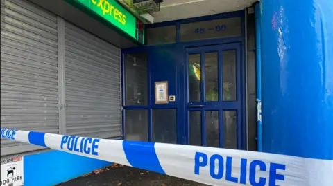 Blue and white police tape in the foreground of a shot of a shuttered shopfront and a door to a block of flats 