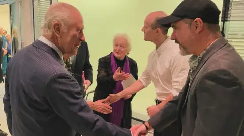 Contributed On the left is King Chales III wearing a navy and white pinstripe suit. He has grey hair, and is holding his hand out shaking it with Steve Young. Steve is wearing a grey suit jacket and is wearing a black baseball hat. 