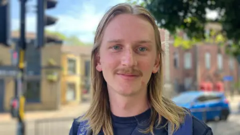 BBC/Elizabeth Baines A man with long blonde hair wears a blue shirt and a blue backpack. He has a moustache. Behind him, a row of a shops are blurred.