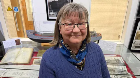 Becki Bowden / BBC A woman wearing a purple jumper, scarf, and glasses. She has short brown hair. She is standing in front a cabinet of memorabilia, including photos, drawings and a model of a ship, and is smiling at the camera.